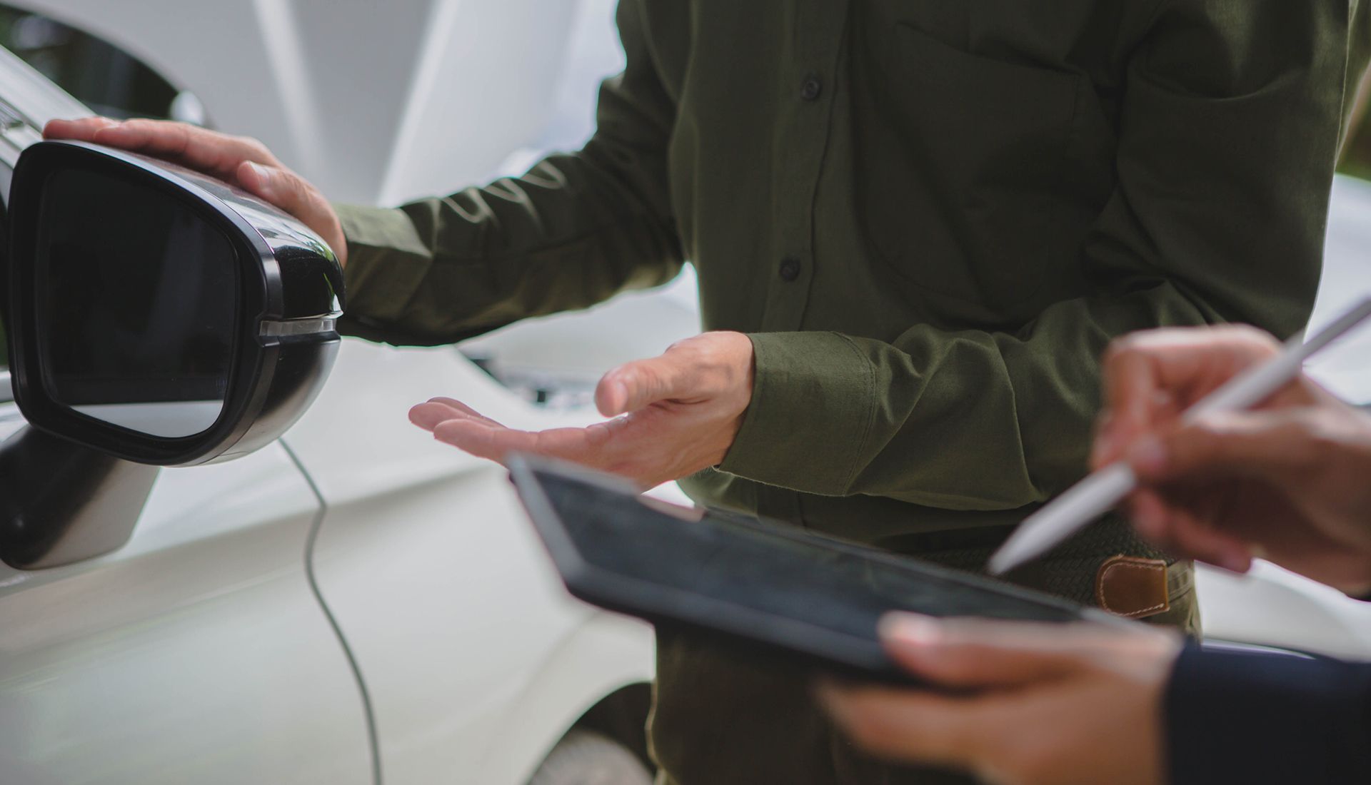 Two people examining a white car; one points to a side mirror while the other holds a tablet and stylus.
