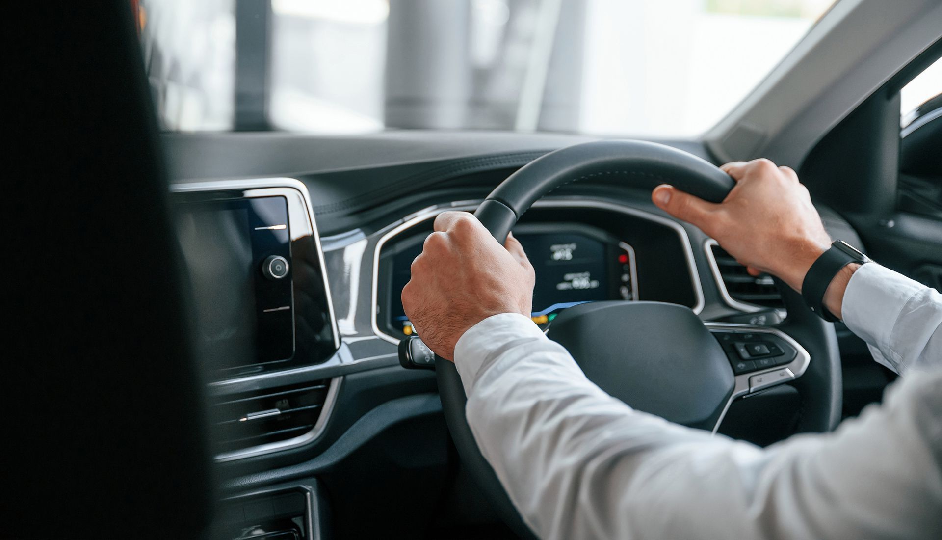 Hands of a person holding a steering wheel in a car. The interior has a digital dashboard and air vents.