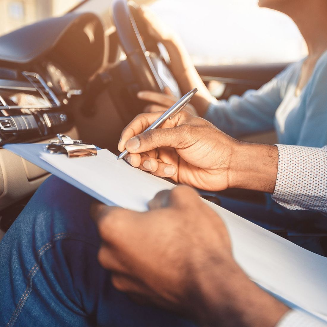 Hands holding a clipboard and pen in a car, writing notes as a person drives, sunlit interior.