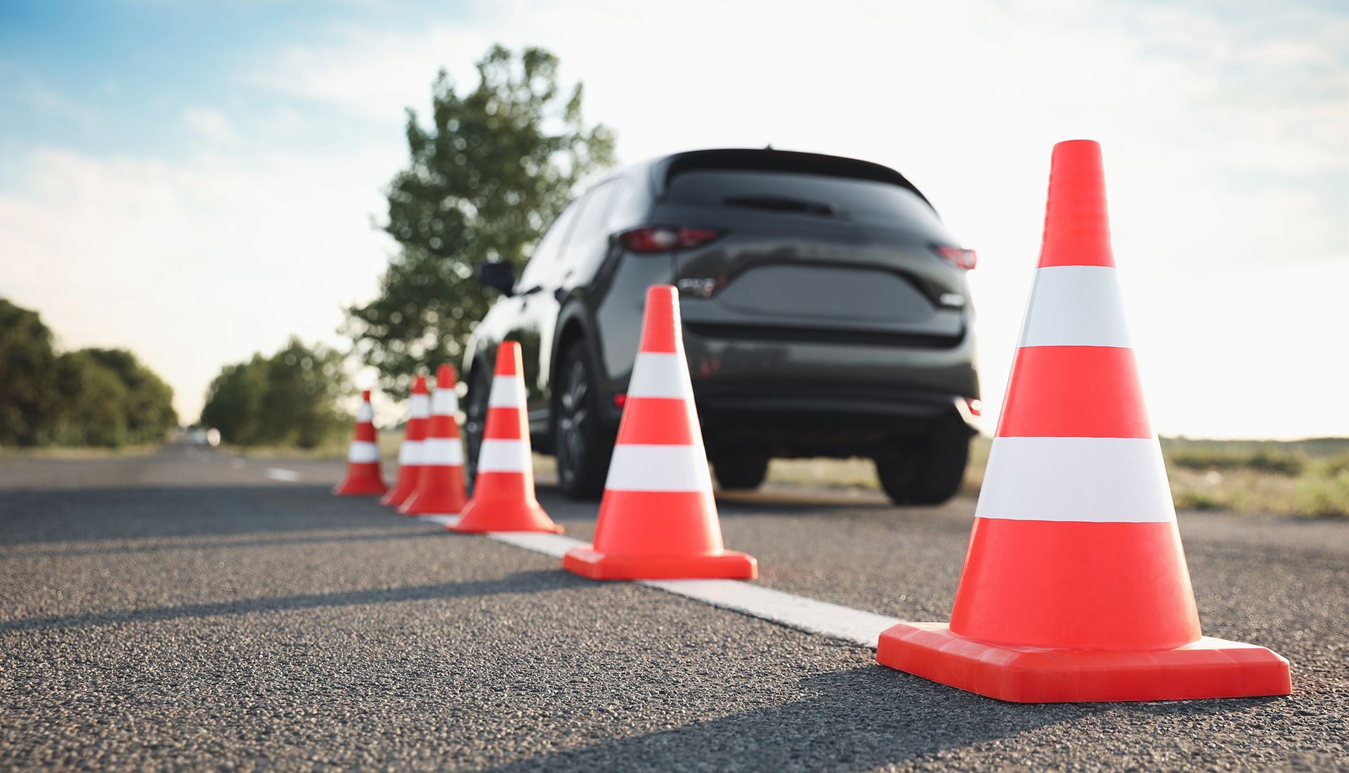 Orange traffic cones line a road, directing traffic around a gray SUV. Sunny outdoor scene.