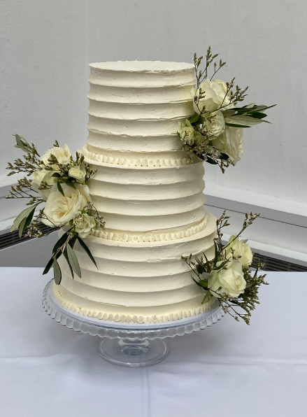 A white wedding cake with white flowers on top is sitting on a glass cake stand.