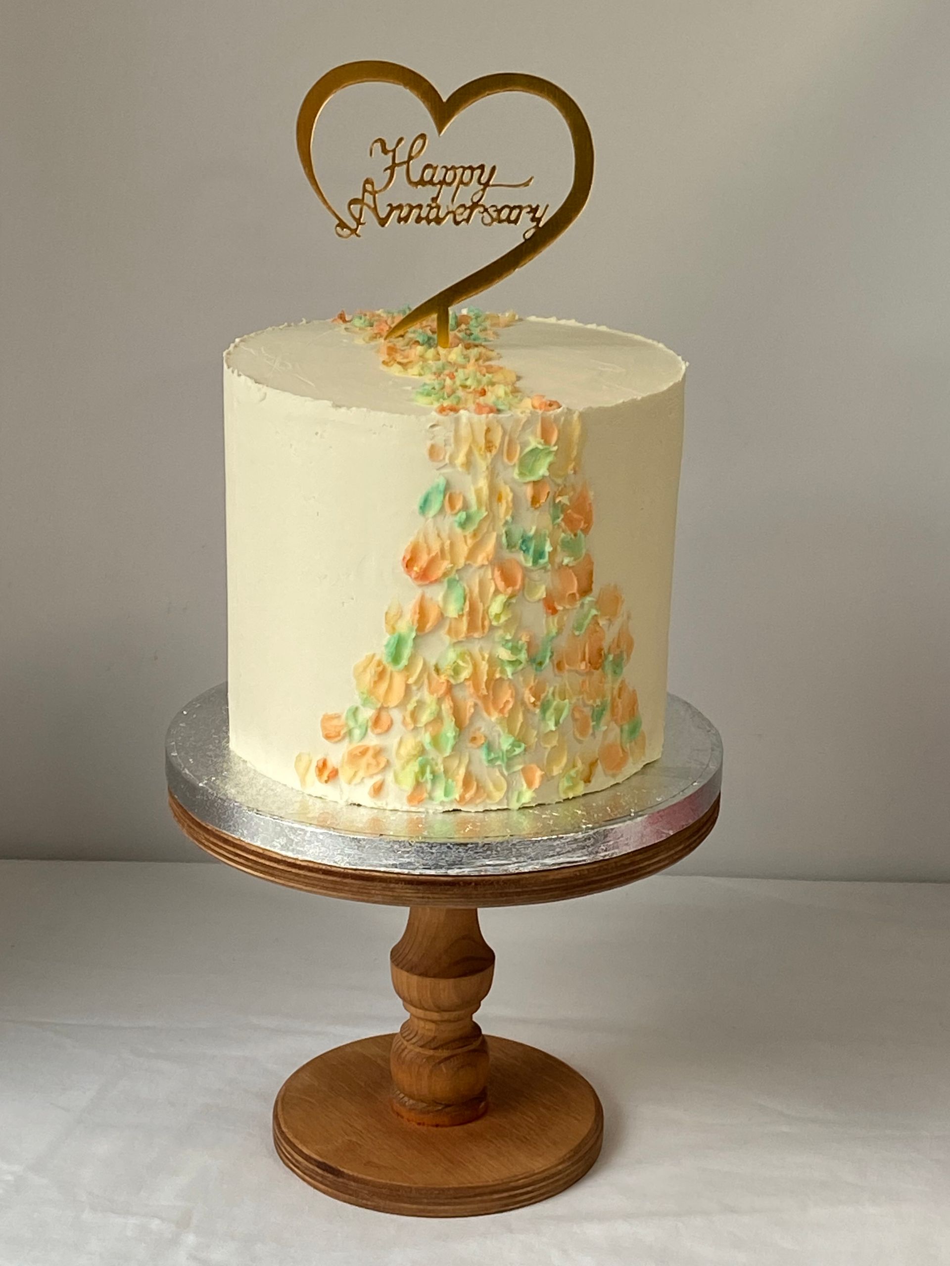 A white cake with a heart shaped topper is on a wooden cake stand.