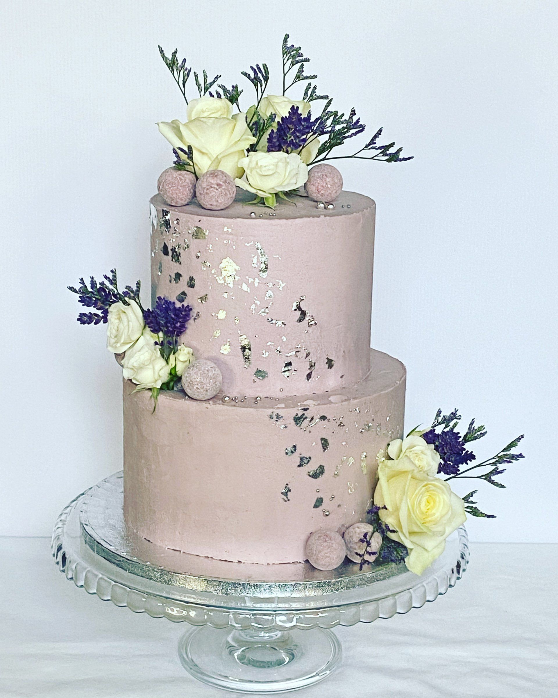 A pink cake with purple and white flowers on top is on a glass cake stand.