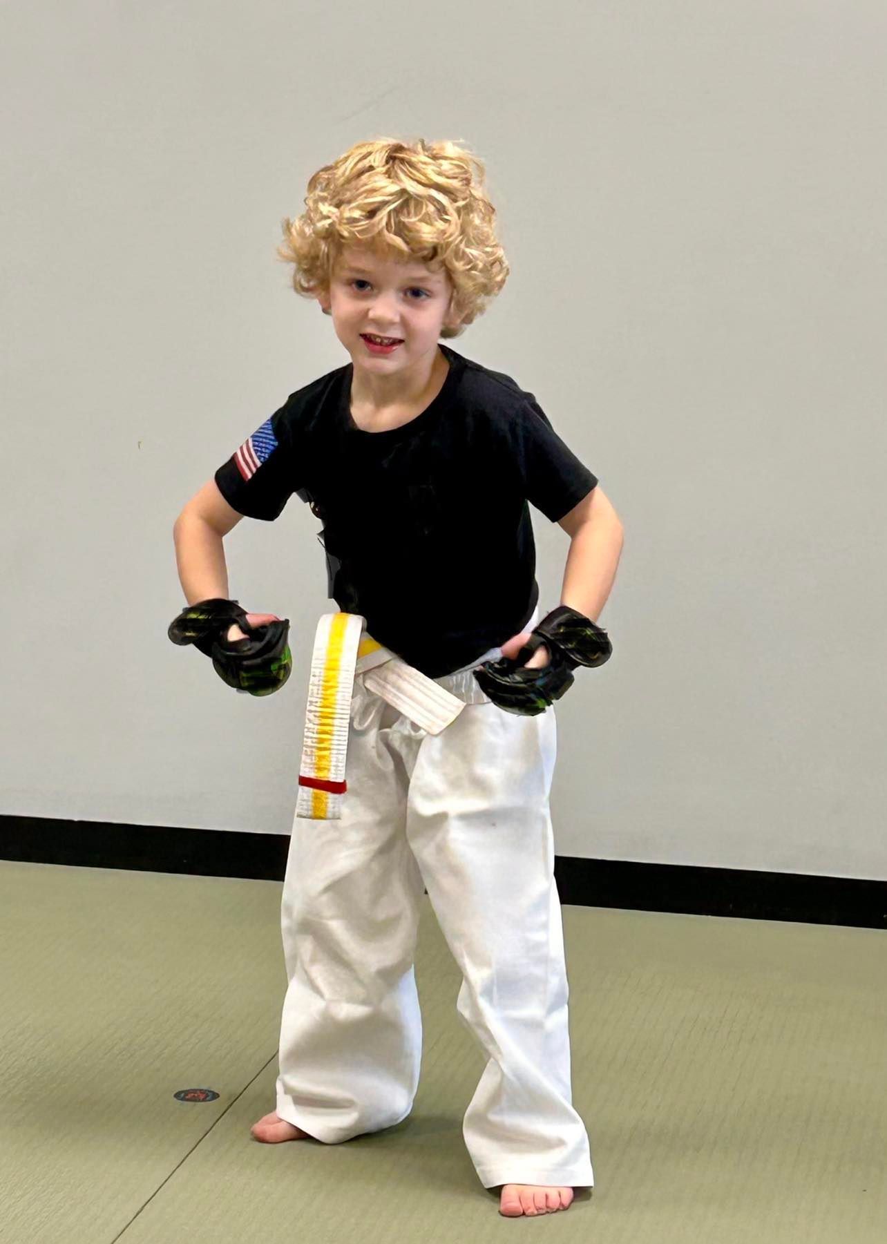 A young boy wearing a black shirt and white pants is standing on a green mat.