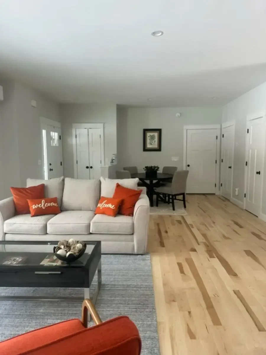 Living room with a beige sofa, orange pillows, and wooden floors. Dining table visible in the background.