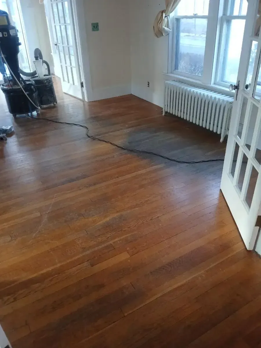 Hardwood floor being refinished; a dark area shows staining. A worker and equipment are visible.