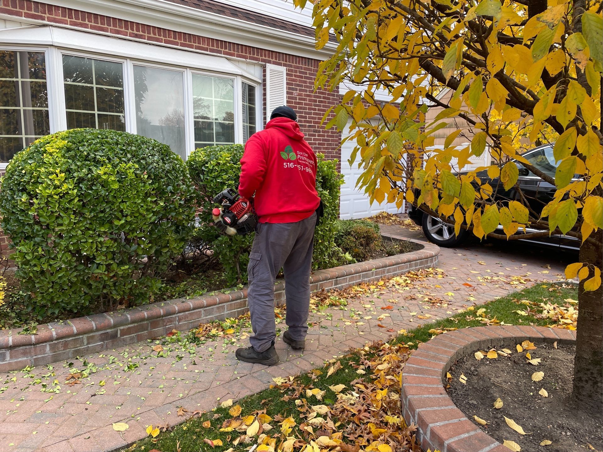 A man in a red hoodie is cutting a bush in front of a house.