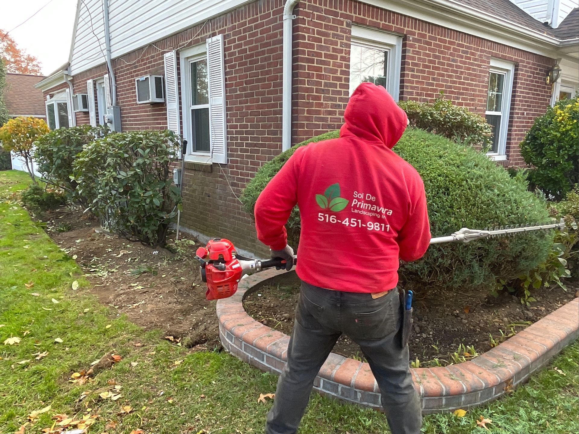A man in a red hoodie is using a lawn mower in front of a brick house.