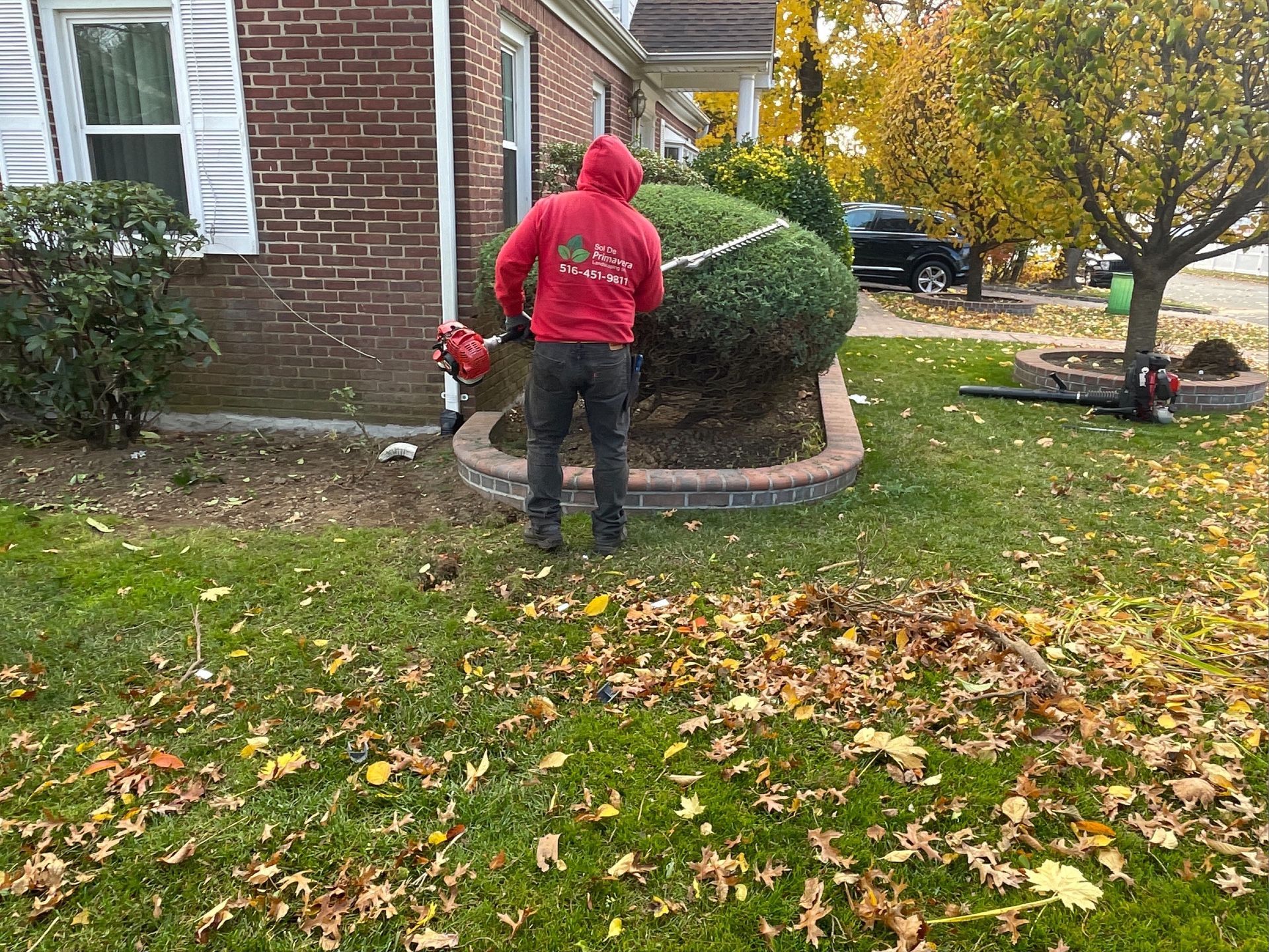 A man in a red hoodie is raking leaves in front of a brick house.