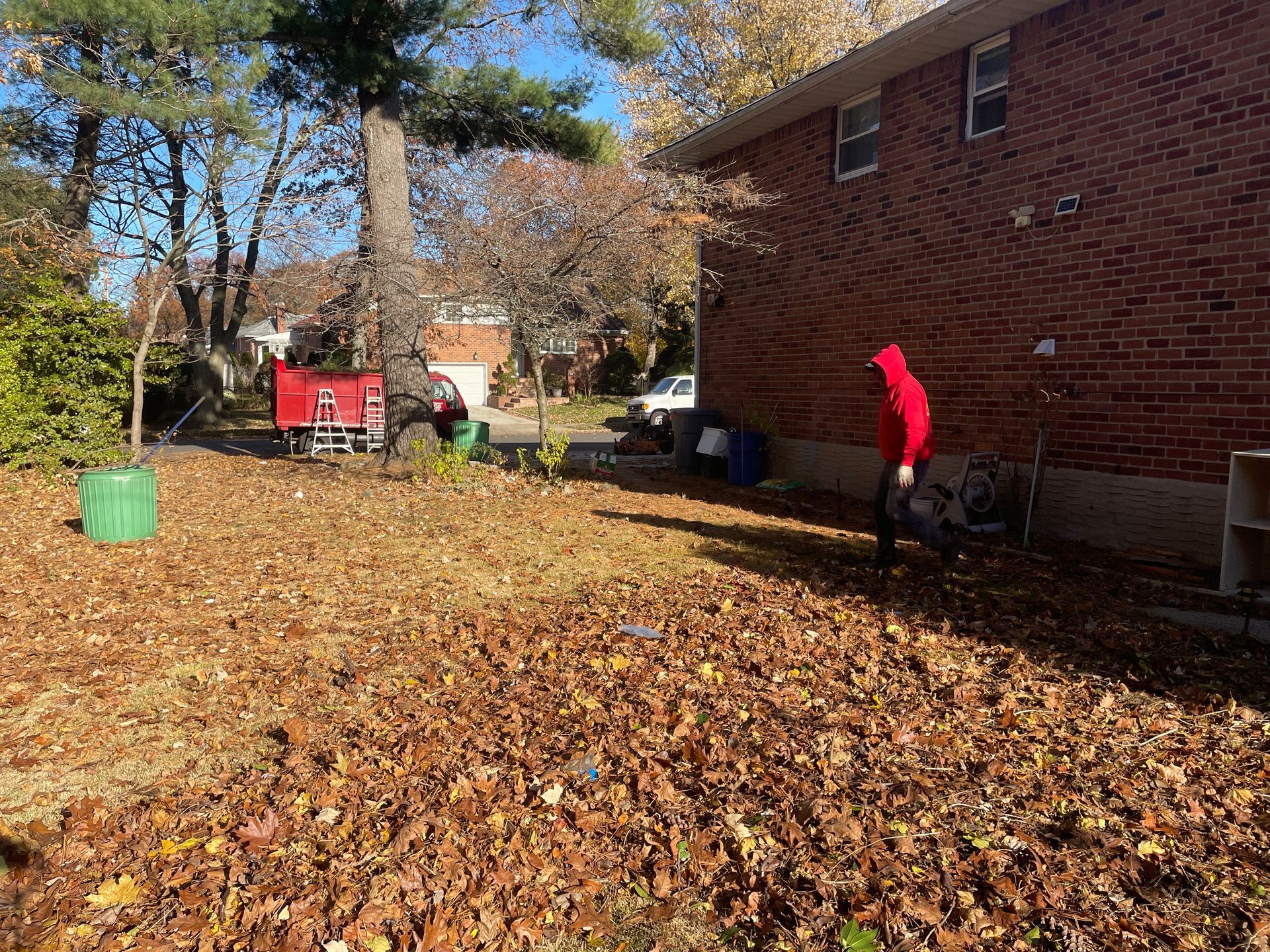 A man in a red jacket is raking leaves in front of a brick house.