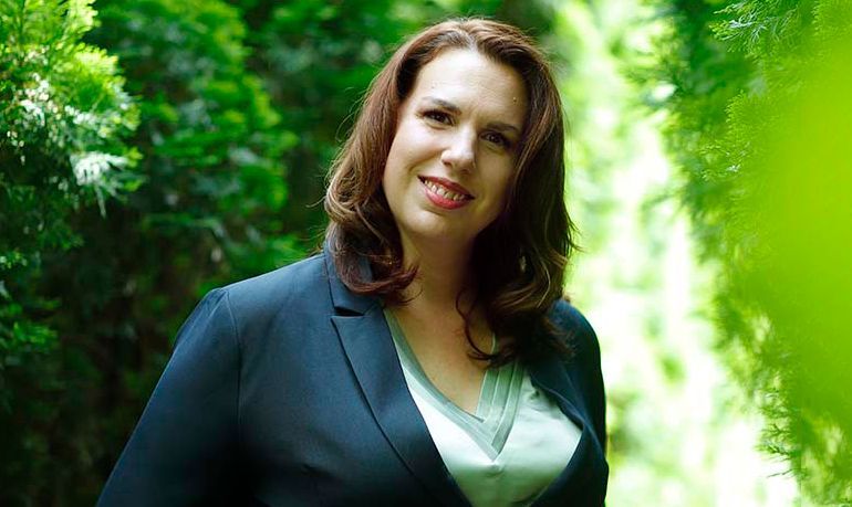 Melanie Jayjack in a blue suit is standing in front of a forest.