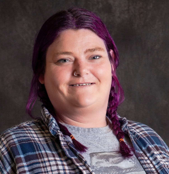 A woman with purple hair is wearing a plaid shirt and smiling.