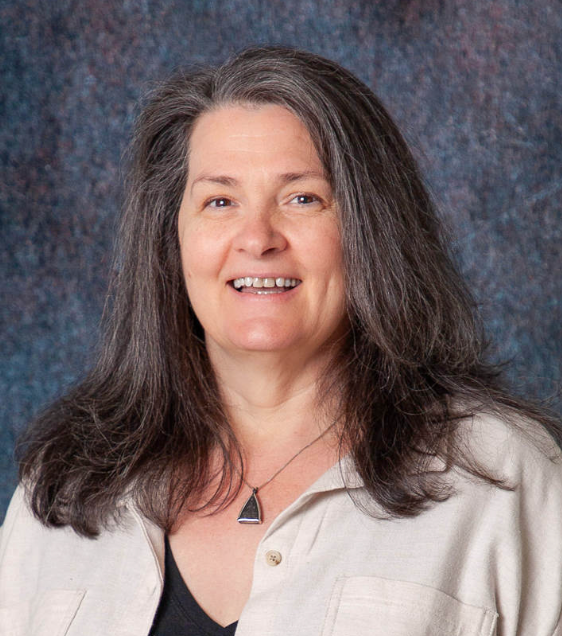 A woman wearing a black jacket and a necklace smiles for the camera