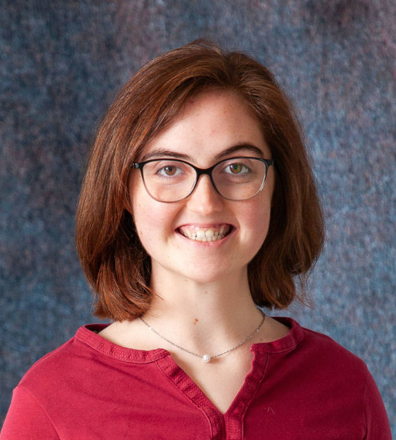 A young woman wearing a blue sweater and a brown shirt smiles for the camera