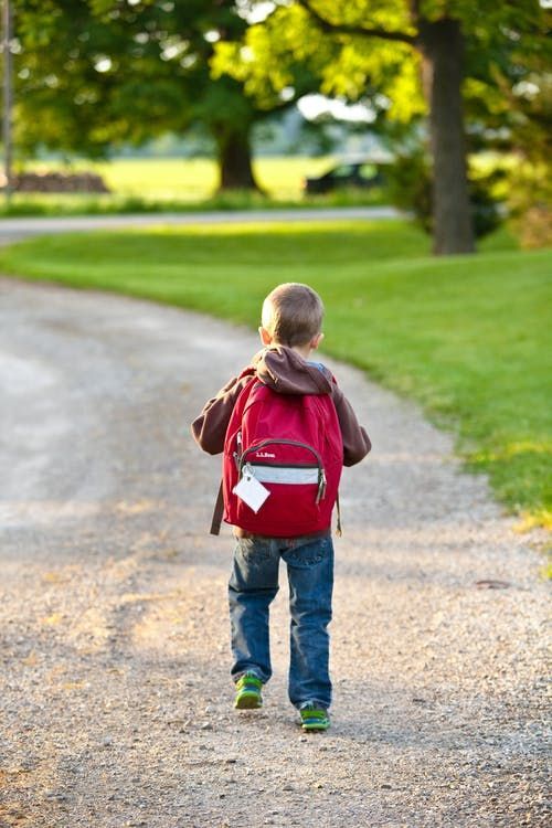 A young boy with a red backpack is walking down a dirt road.