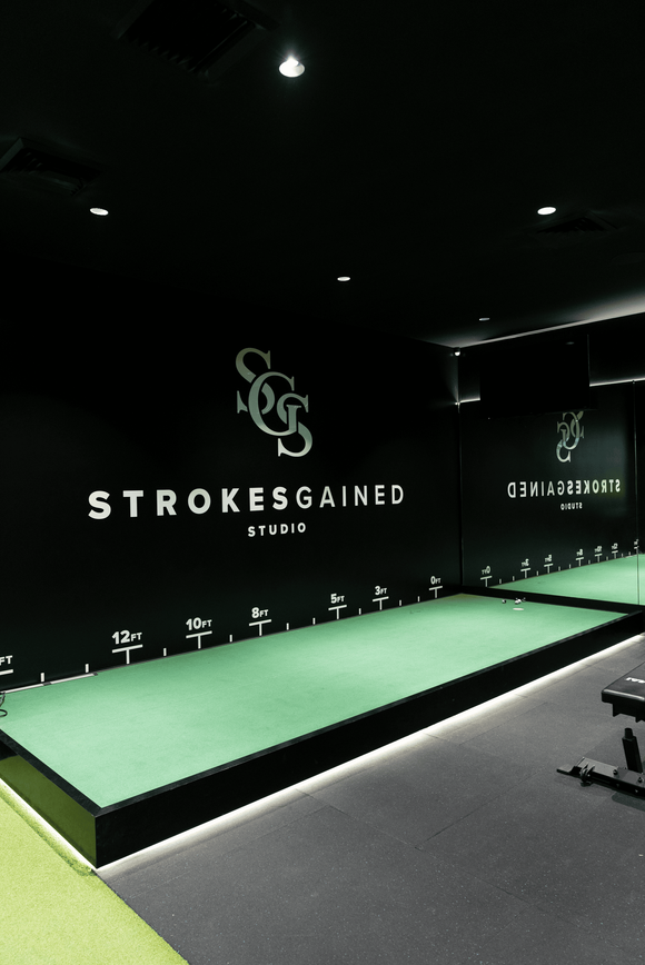 Indoor golf studio with putting green, mirrored wall, kettlebells, and golf simulator in the background.
