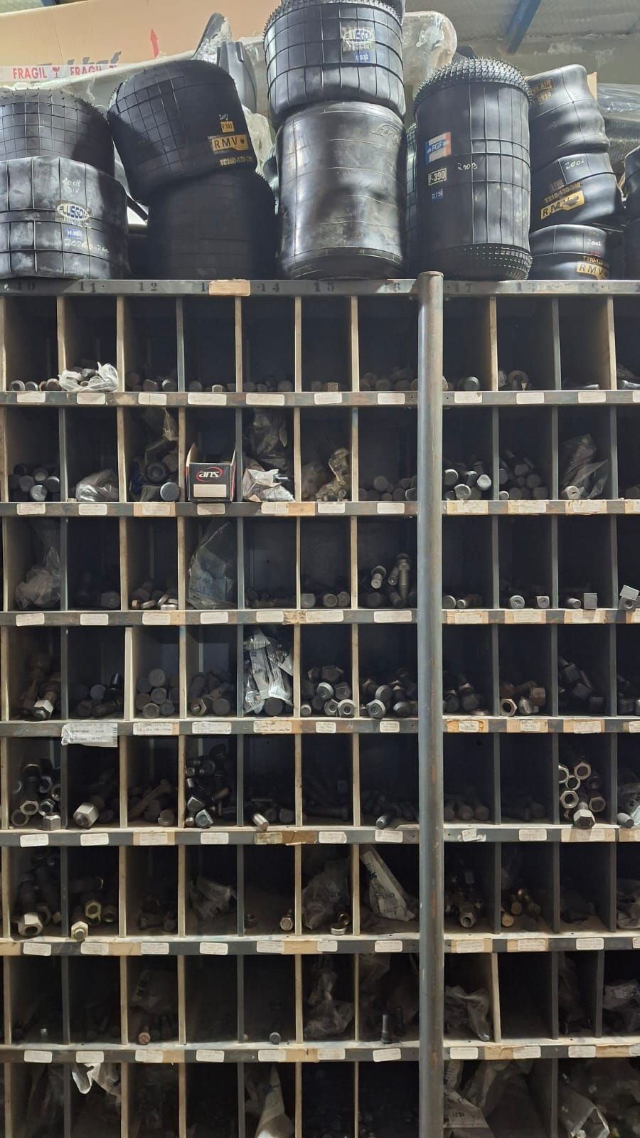 Estantería llena de piezas pequeñas, probablemente hardware, debajo de grandes objetos cilíndricos negros.