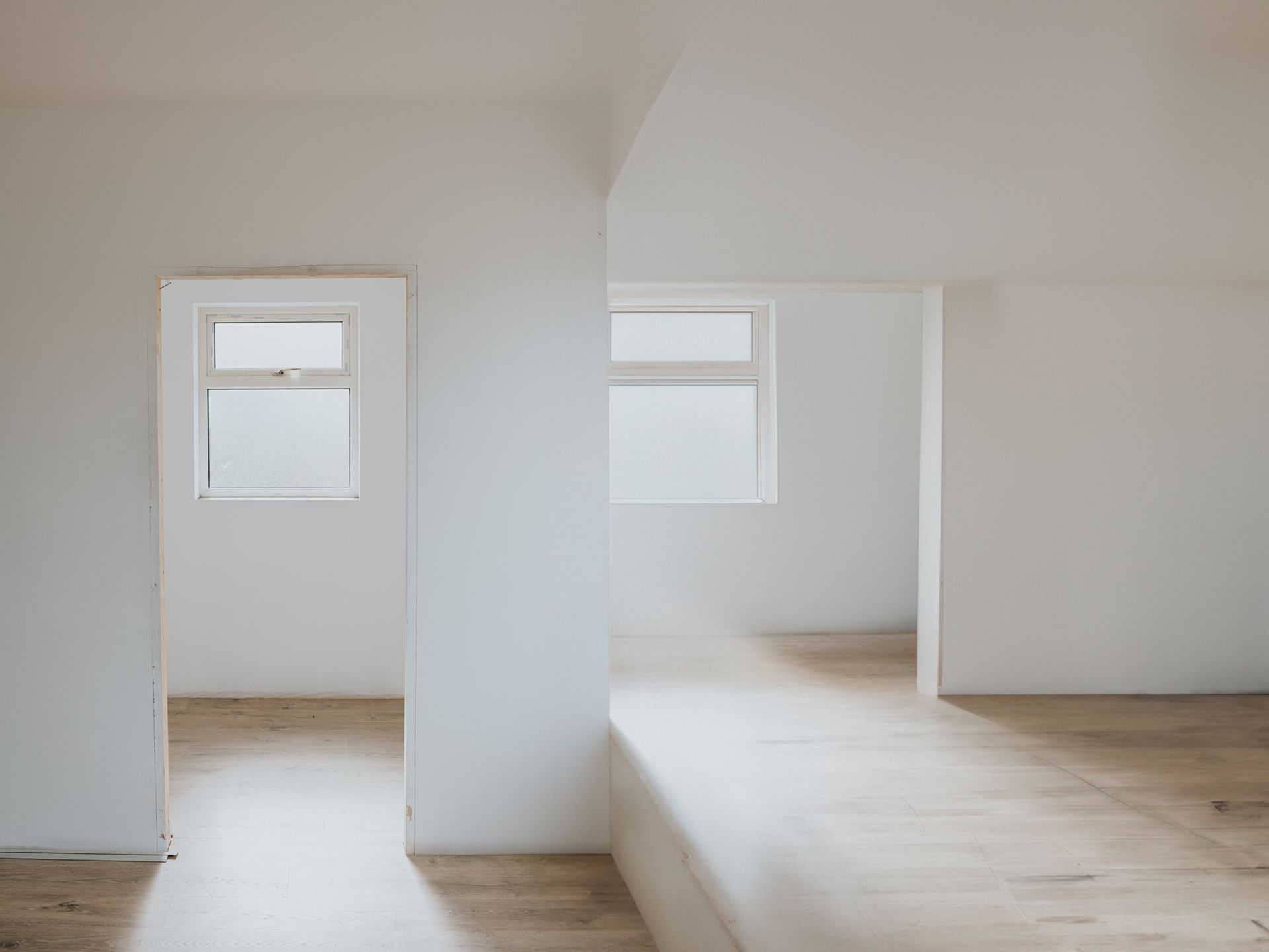 Minimalist white room with wooden floors and two windows providing natural light.