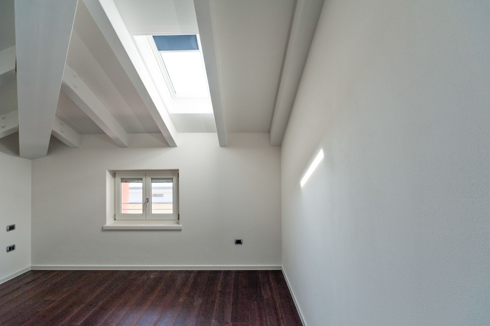 Empty room with white walls, a dark wooden floor, and a skylight above a small window. Empty room with white walls, a dark wooden floor, and a skylight above a small window.