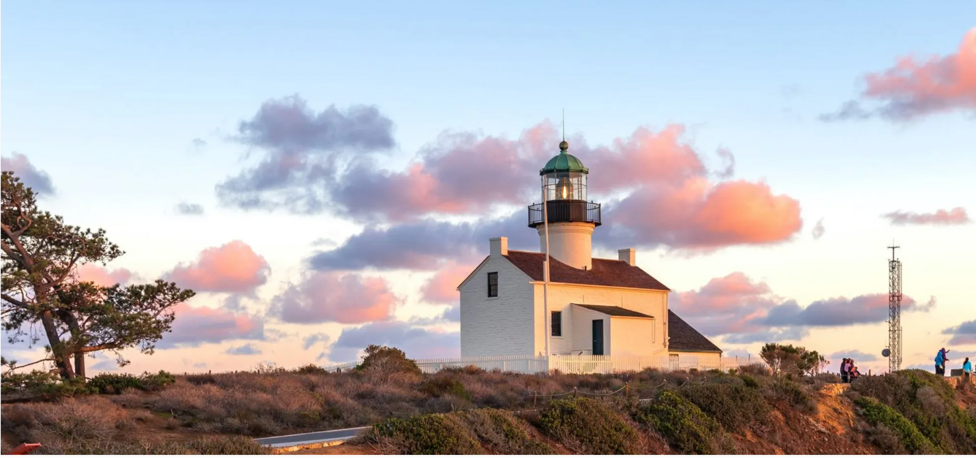 Point Loma Lighthouse on a cliff at sunset with pink and gray clouds in the sky.