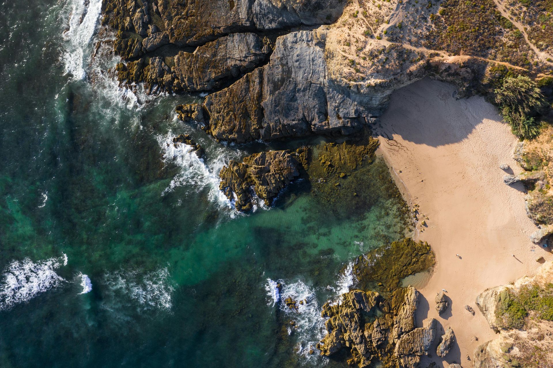 An aerial view of a sandy beach cove meeting rocky cliffs and turquoise ocean waves crashing against the shore.