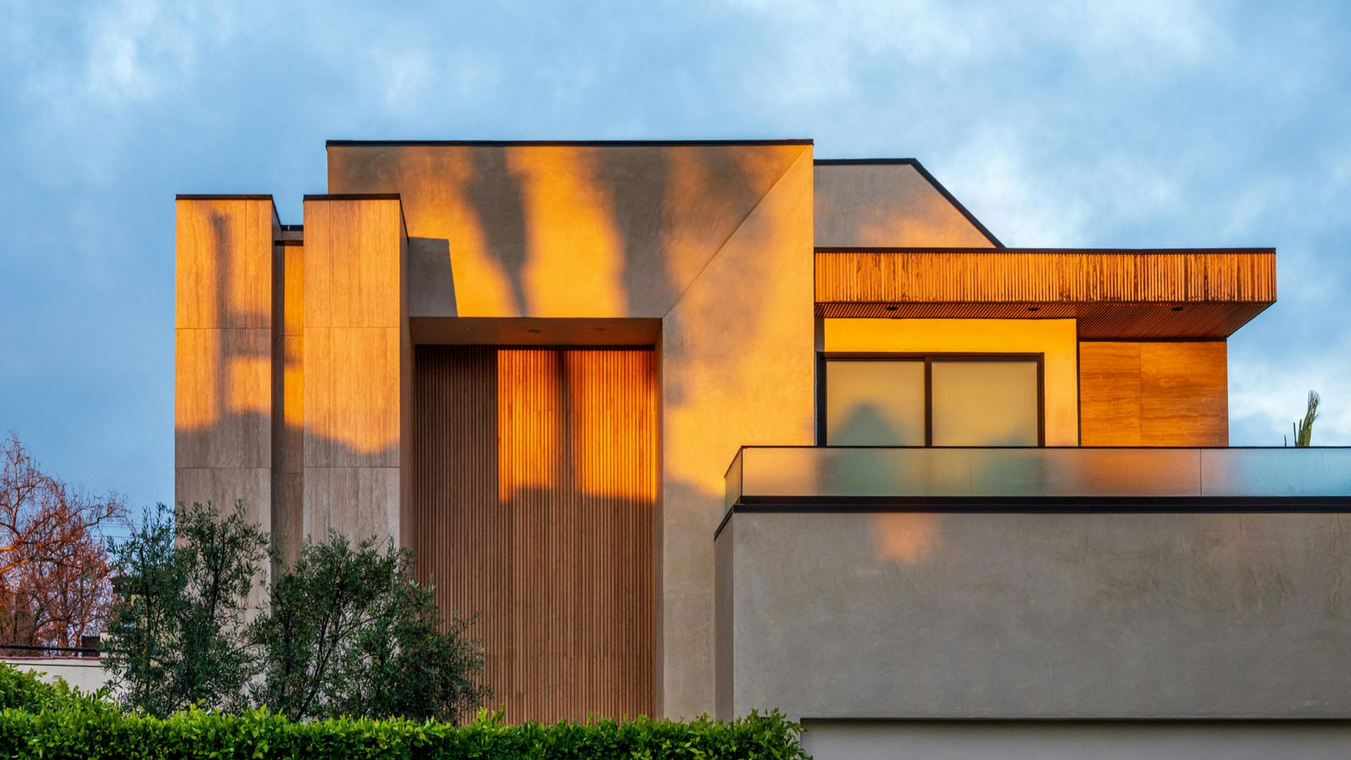 Modern home exterior with warm sunset lighting casting tree shadows on beige walls, wooden accents, and a glass balcony.