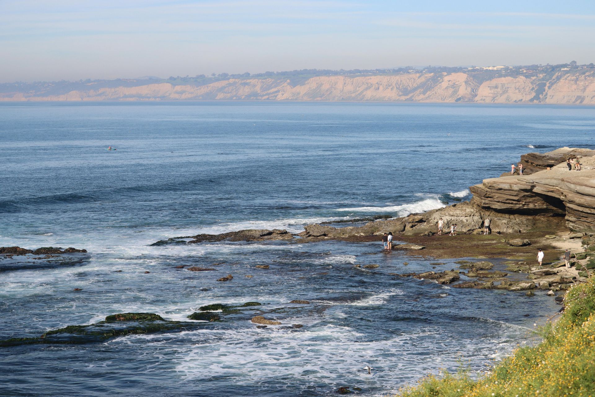 Rocky shoreline with calm blue water and distant cliffs under a hazy sky