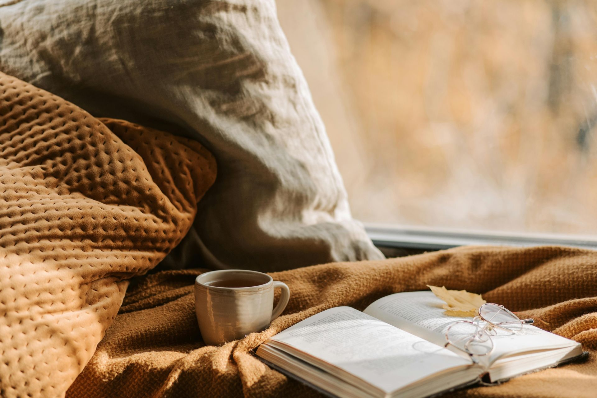 Open book with yellow leaf, cup of tea, and cozy blanket by a window.
