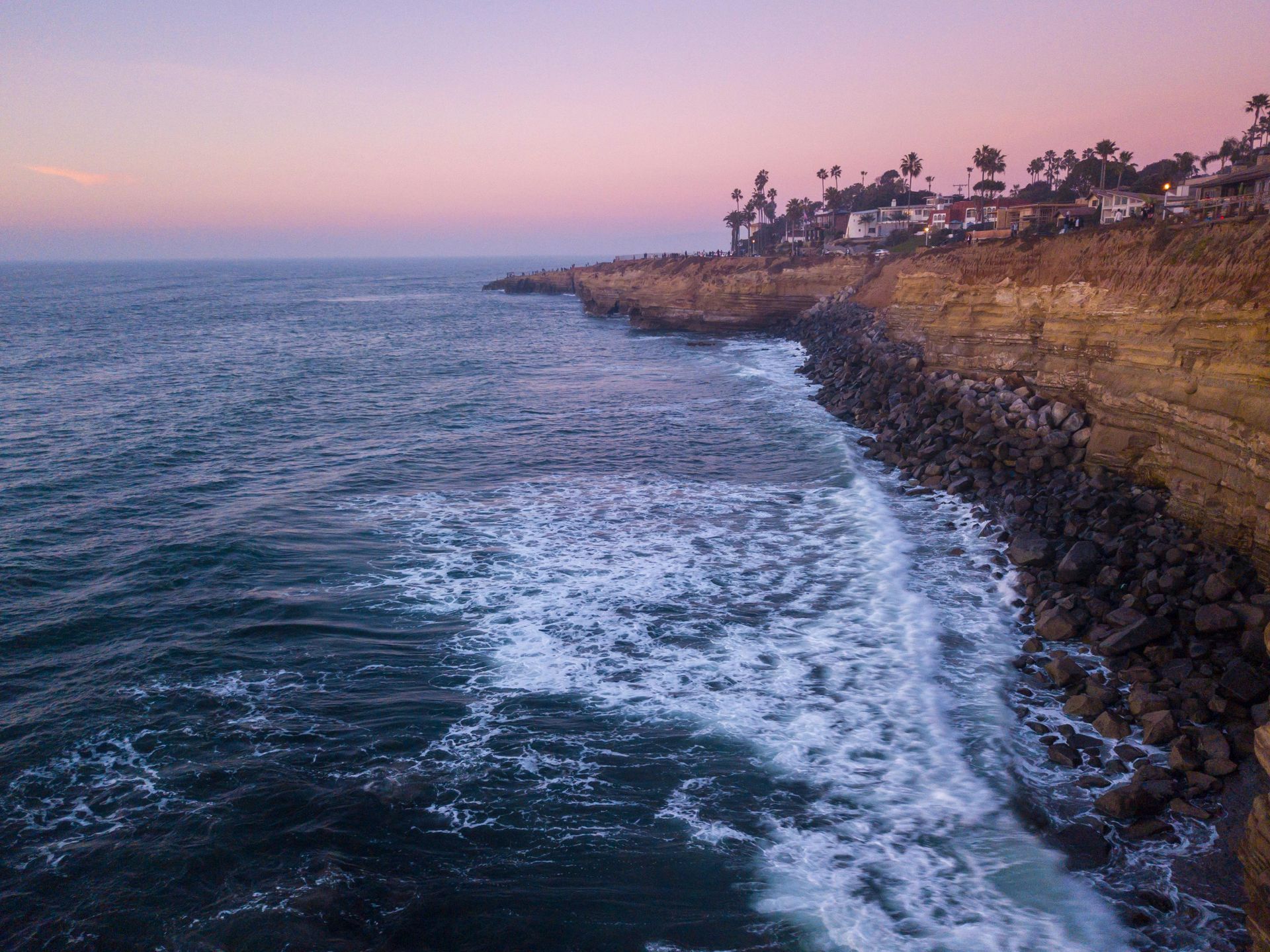 Ocean waves crashing against a rocky shoreline at sunset, with buildings visible on a cliffside in Point Loma 92106