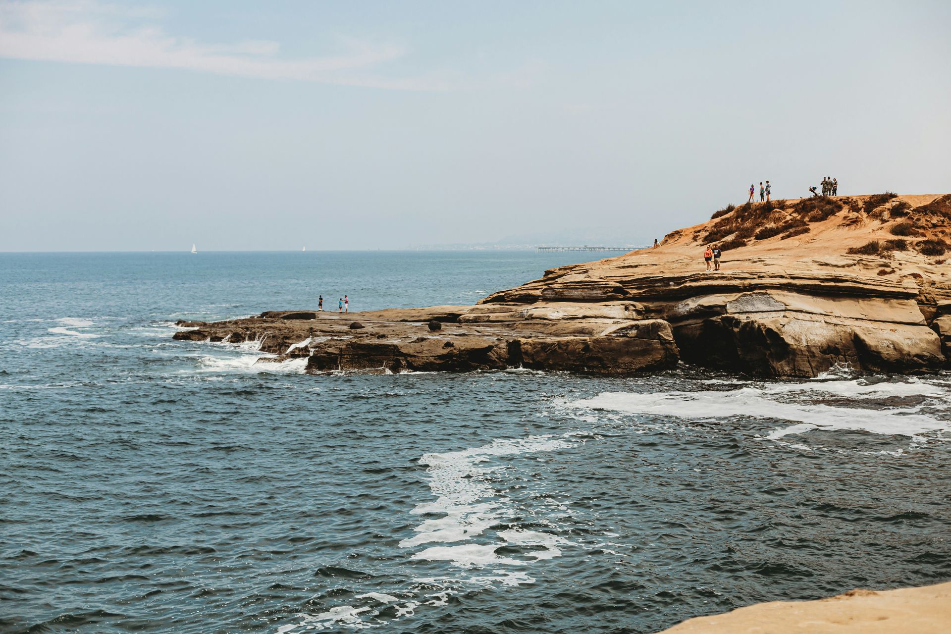 Rocky coastline with ocean waves and people on a sandy bluff under a blue sky in Point Loma