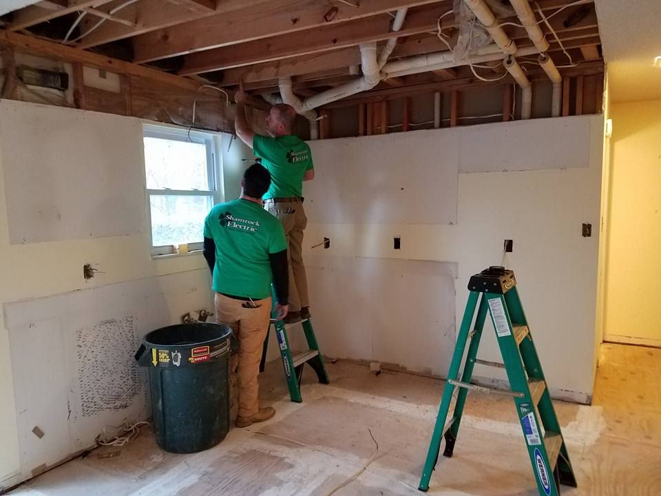 Two workers in green shirts renovate a kitchen, one on a ladder, plumbing exposed.