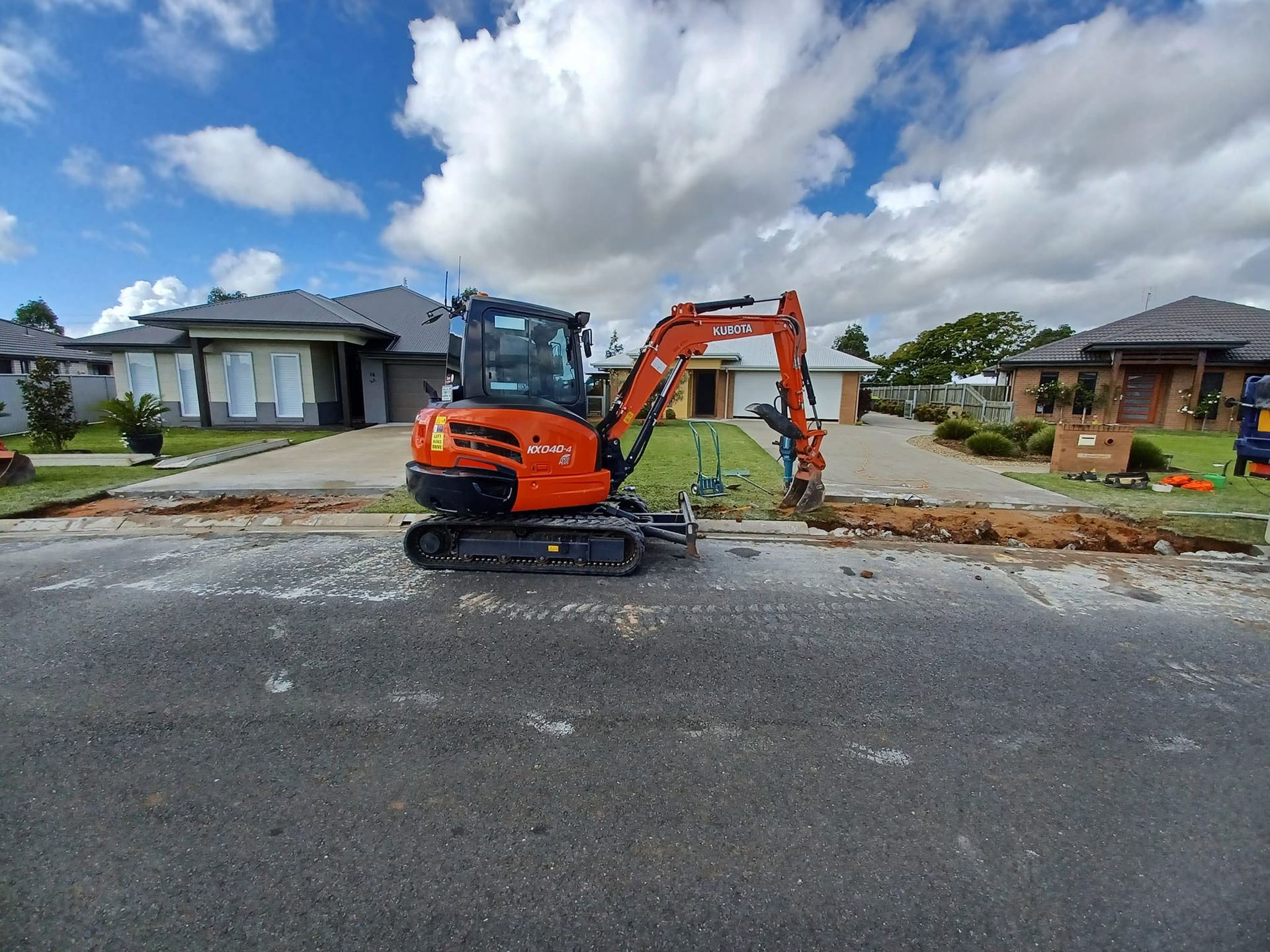 An orange mini excavator digs a trench in front of houses — Brocksolid Concrete in Pillar Valley, NSW