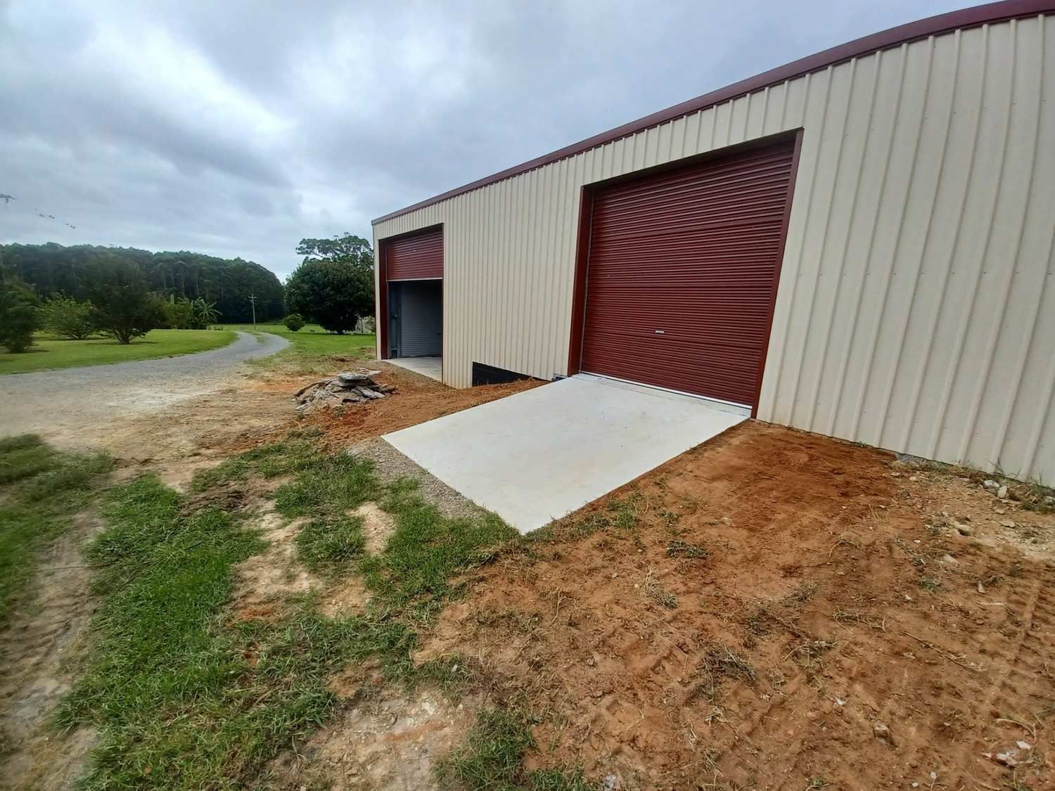 A Large Metal Building With a Red Garage Door is Sitting on Top of a Dirt Field — Brocksolid Concrete in Pillar Valley, NSW