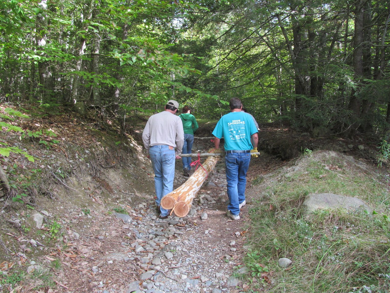 Two men are carrying a large log down a dirt path in the woods.