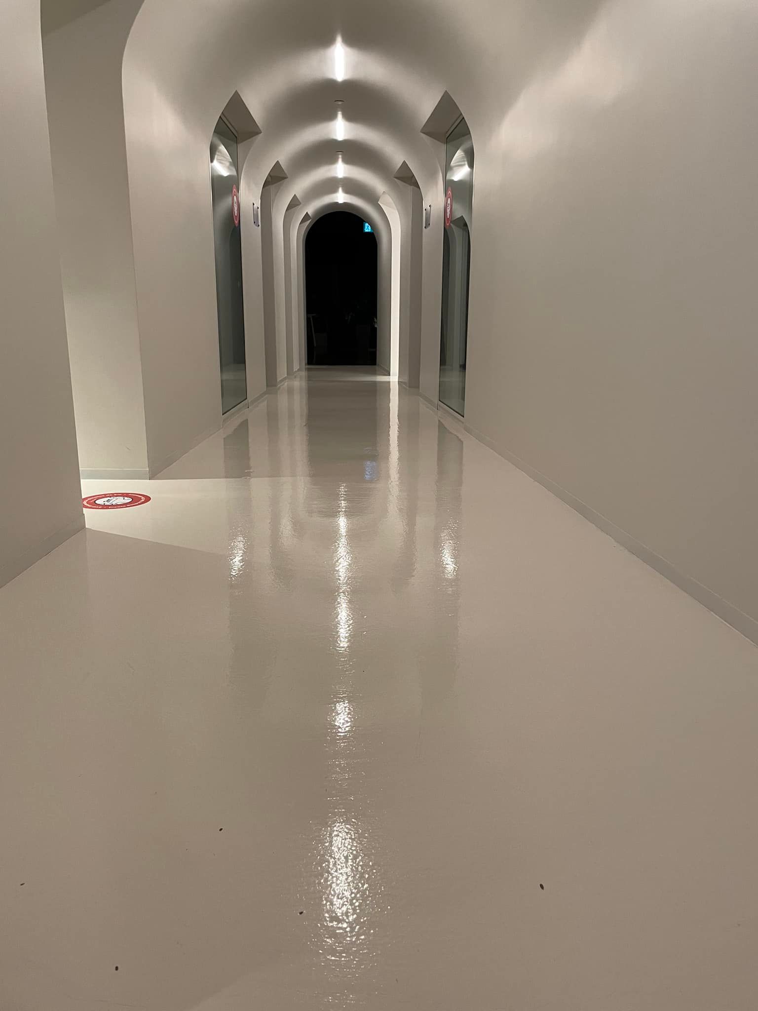 Long couloir blanc au plafond voûté et au sol réfléchissant, menant à un fond sombre.