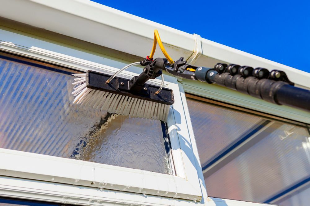 Un laveur de vitres utilise une brosse allongée pour nettoyer une fenêtre, ciel bleu en arrière-plan.
