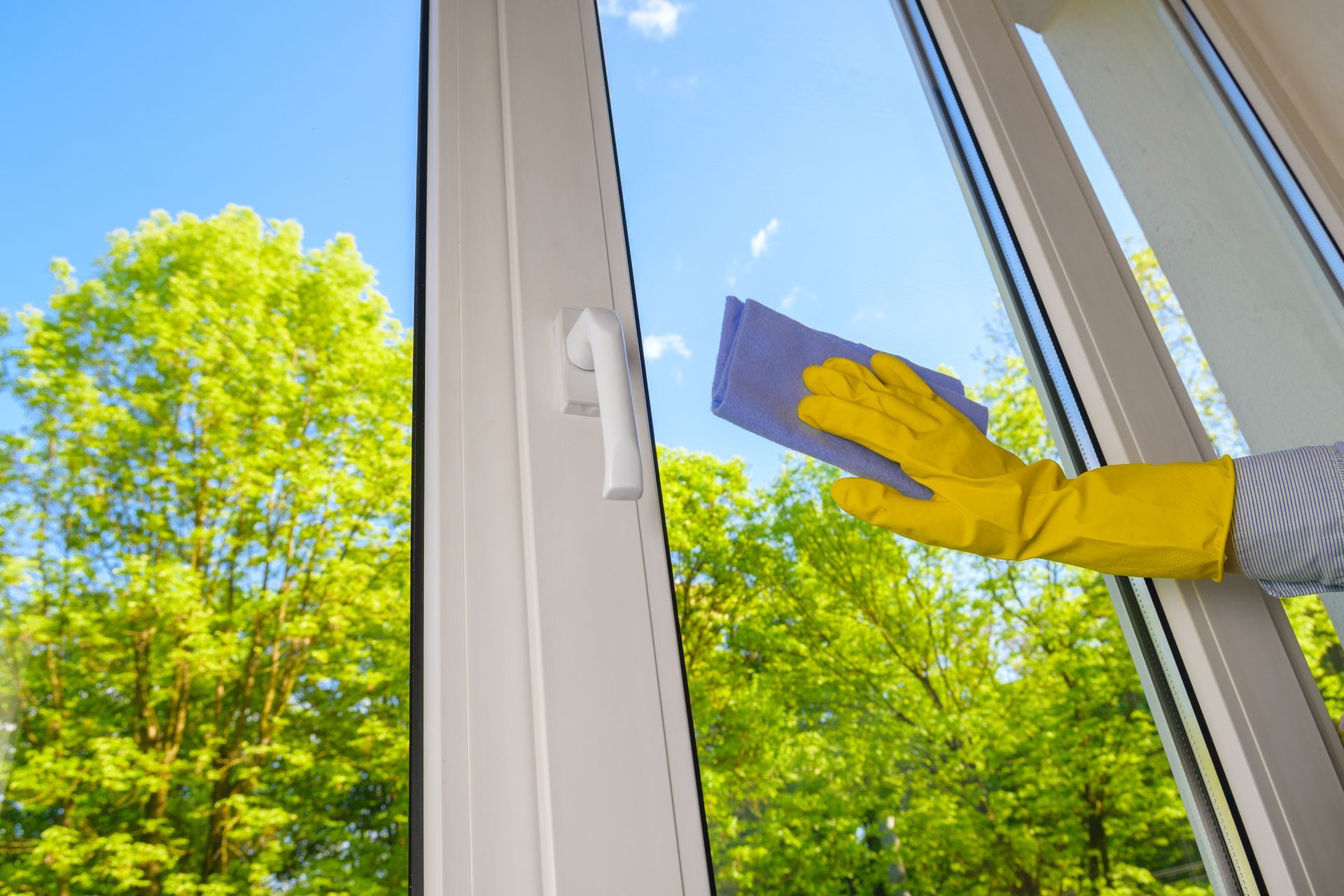 Une personne portant un gant jaune essuie une fenêtre donnant sur des arbres verts et un ciel bleu.