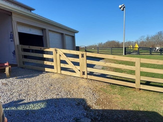 Wooden gate and fence leading from a gravel driveway to a grassy field next to a light-colored building.