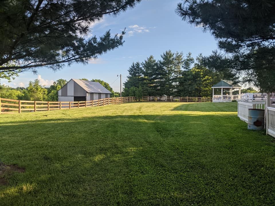 A grassy field with a barn, fence, and gazebo under a blue sky, framed by trees.