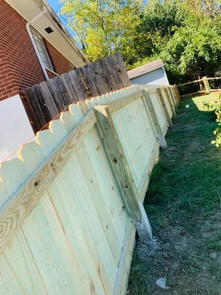 Wooden fence with scalloped top along a brick building and green grass.