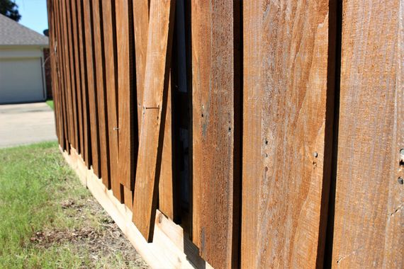 Close-up of weathered, rusty brown wood siding on a building's exterior, with green grass and a garage in the background.