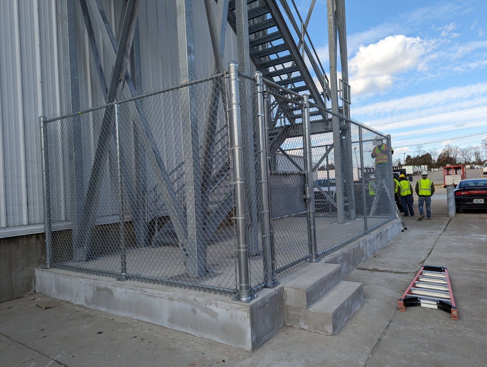 Chain link fence encloses stairs leading up to a metal structure. Two people in vests stand nearby.