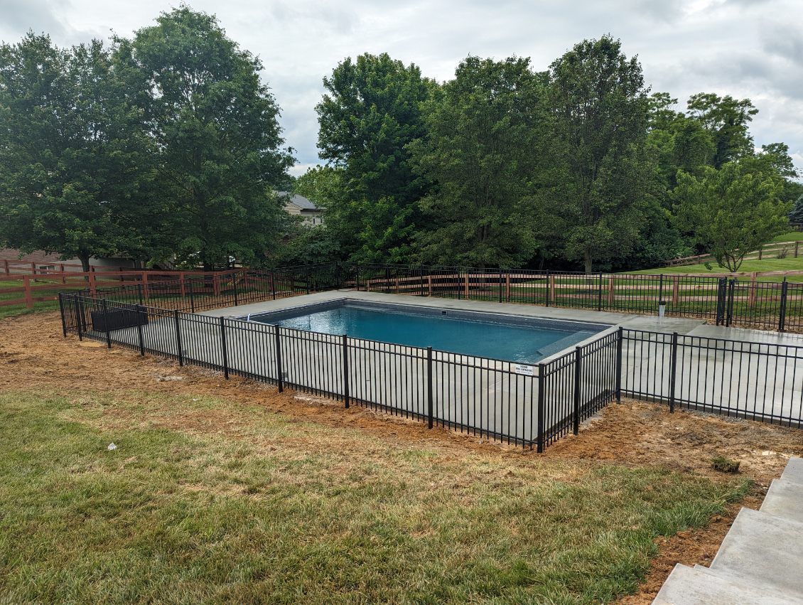 Above-ground rectangular pool surrounded by a fence and mulch, backed by trees and a cloudy sky.