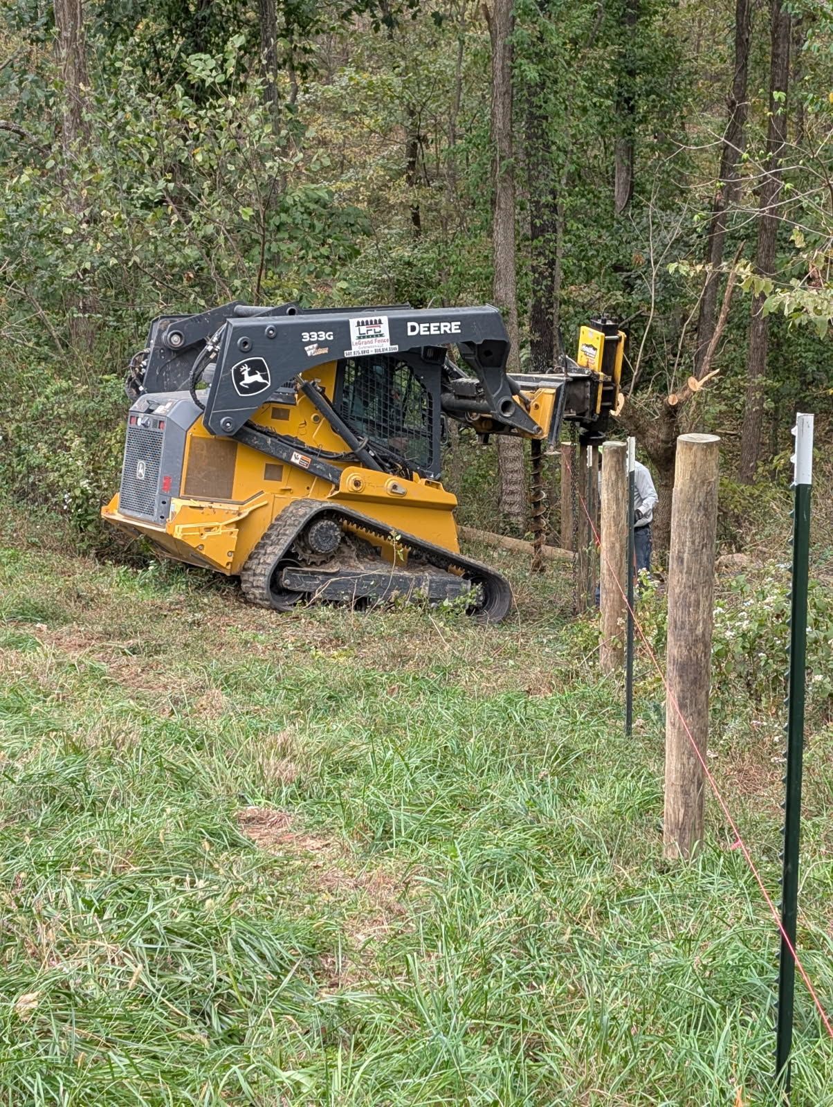 Yellow John Deere skid steer installing fence posts in a grassy area near trees.