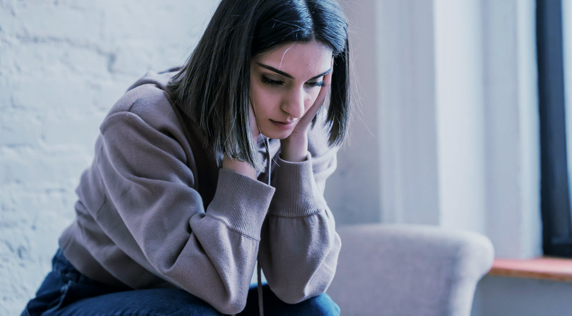 Woman in a pink hoodie, looking down with a sad expression, seated indoors near a window.