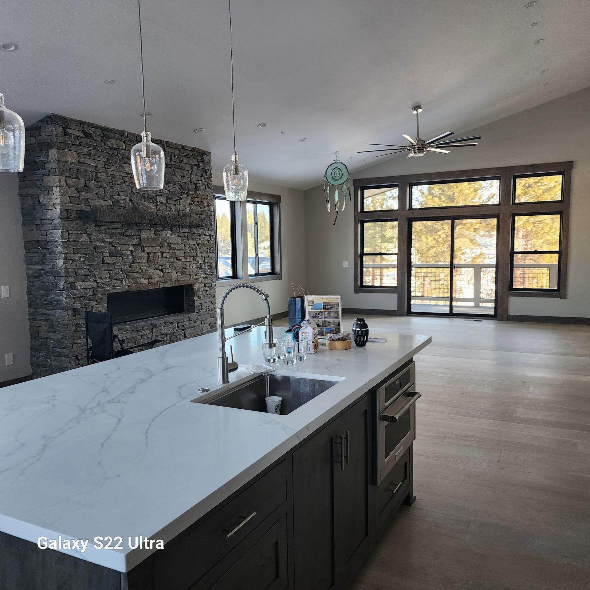 Open kitchen with marble island, dark cabinets, stone fireplace, and large windows overlooking trees.