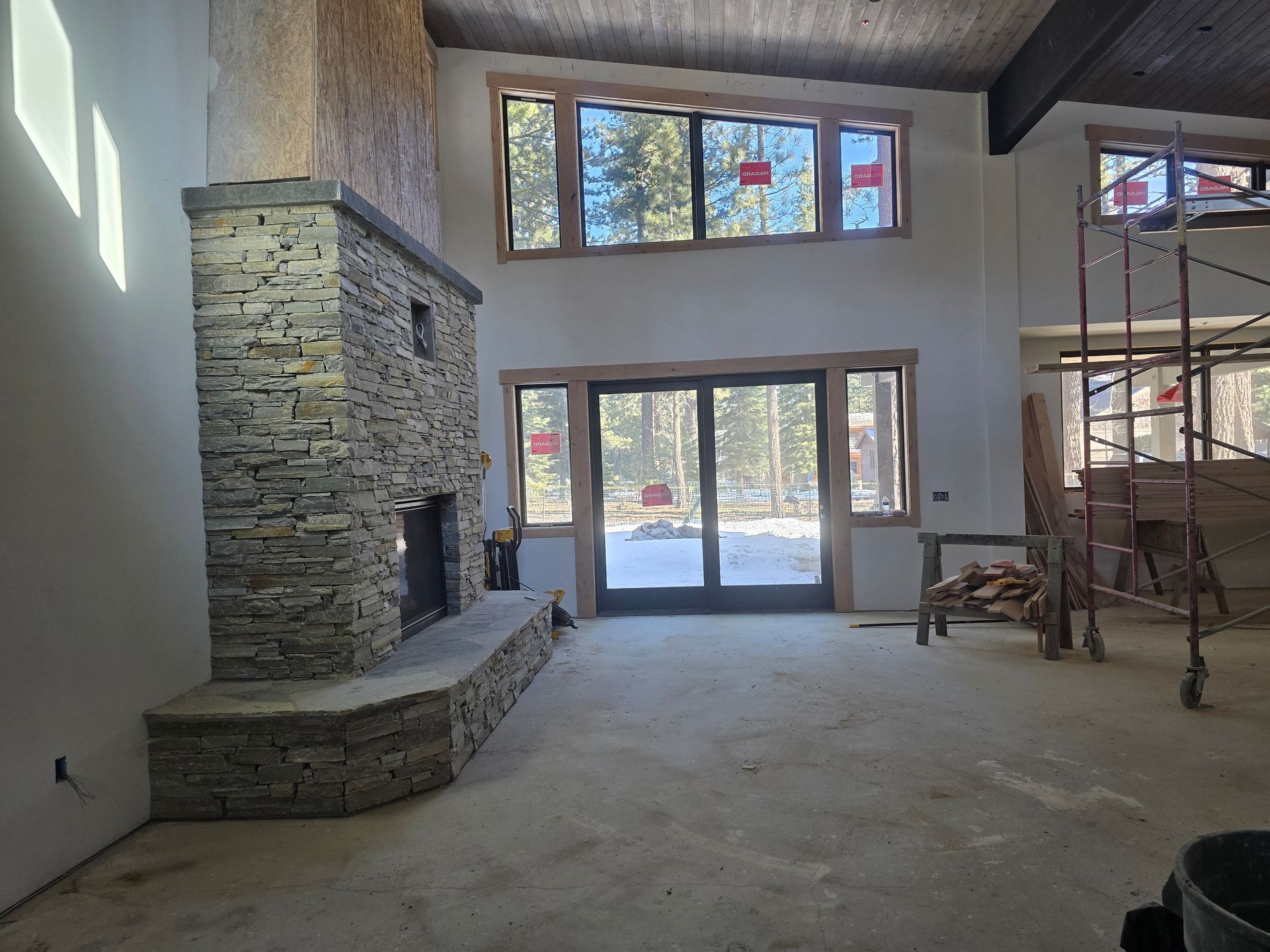 Unfinished living room with a stone fireplace, high windows, and glass doors leading outside.