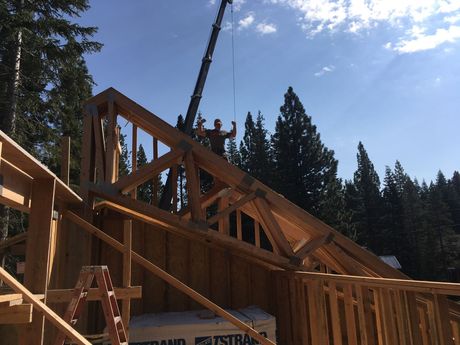 Wooden house frame under construction with a crane lifting roof trusses against a blue sky