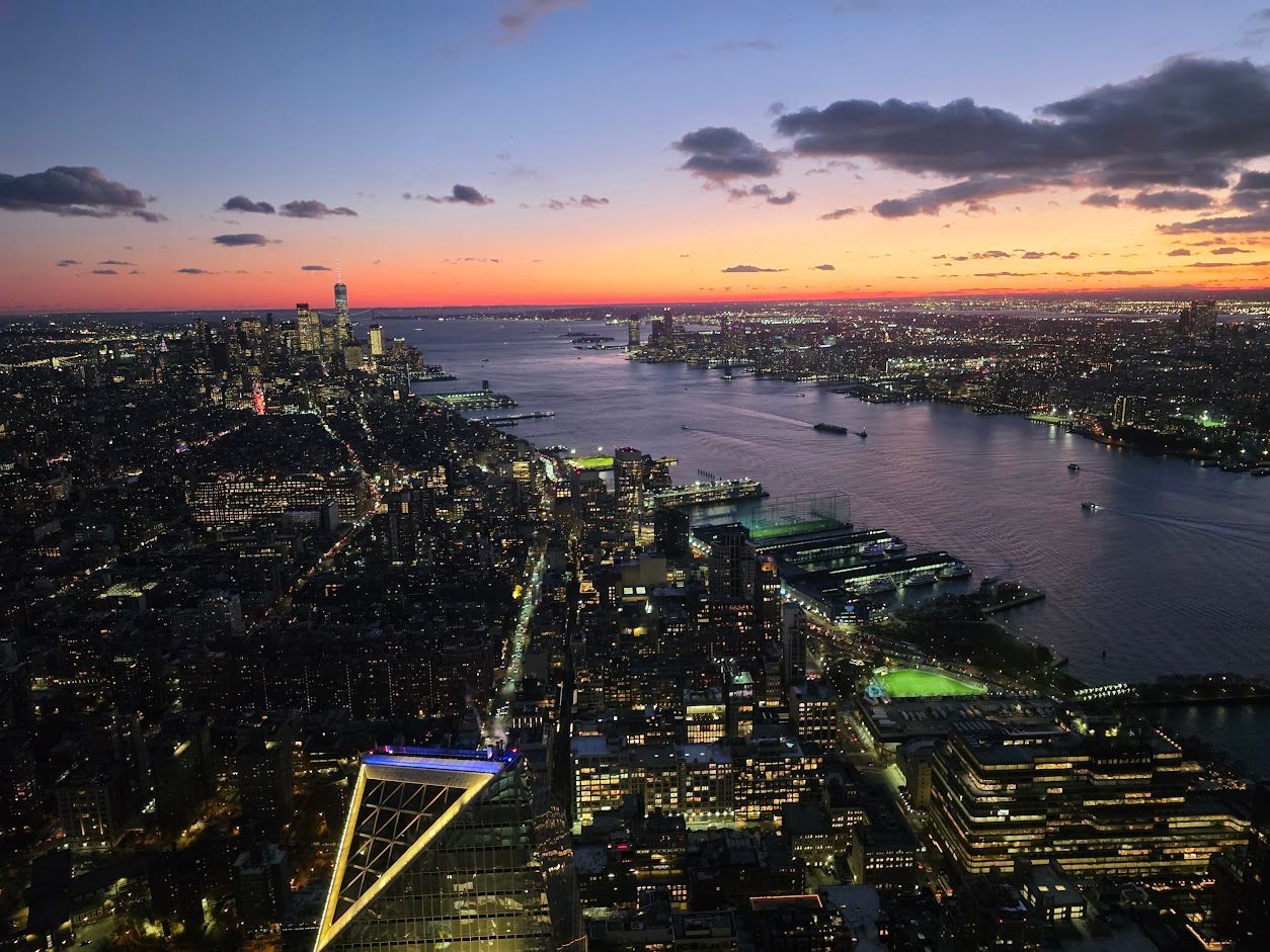 Nighttime aerial view of New York City skyline and harbor at dusk, with city lights and colorful sky.