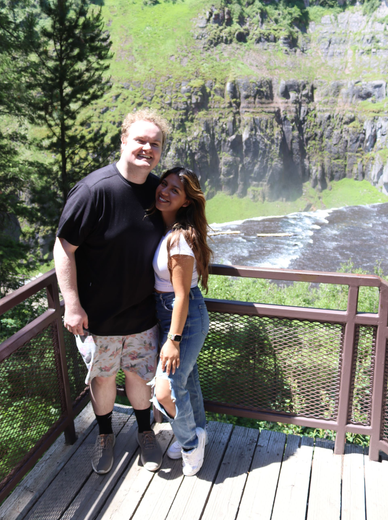 Couple poses on a wooden deck overlooking a waterfall, smiling; sunny day.
